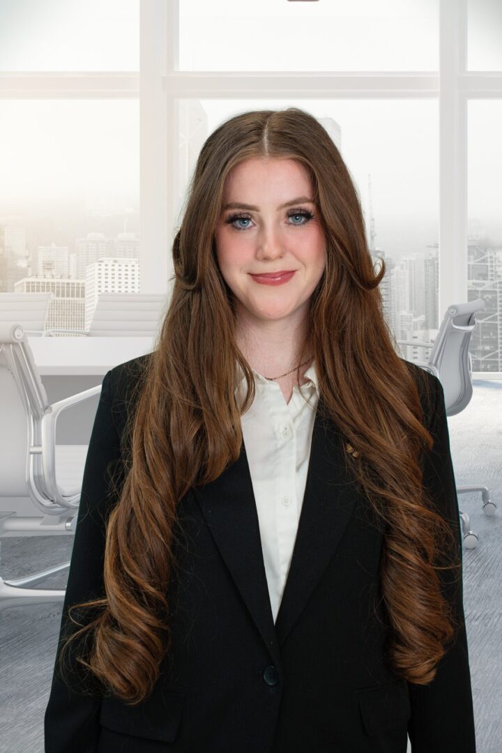 A young woman with long brown hair in business attire stands in a modern office.